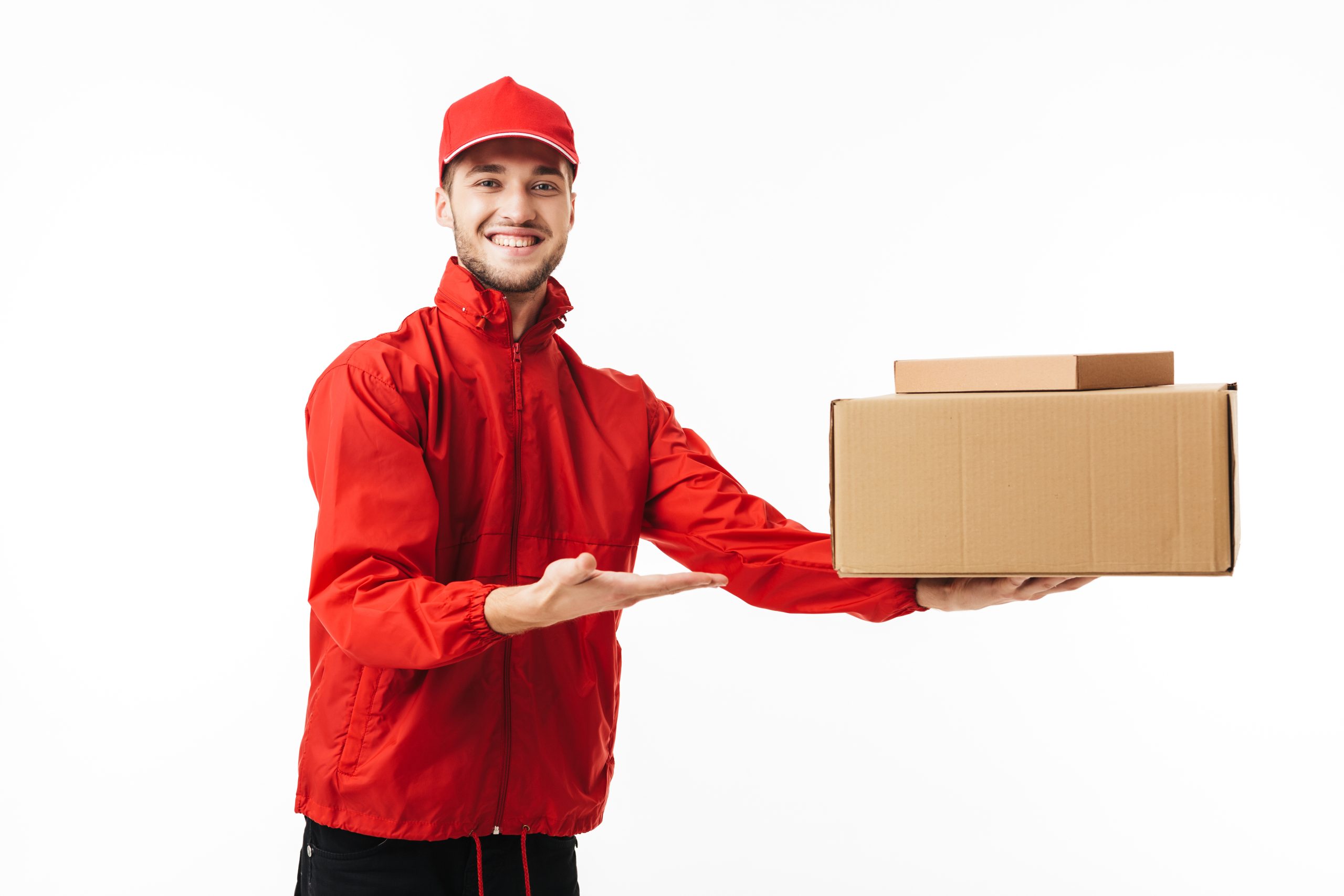 Young joyful delivery man in red cap and jacket happily looking in camera giving package to customer over white background